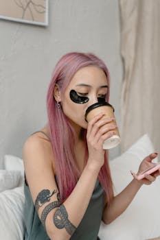 Asian woman with pink hair enjoying a coffee while wearing eye patches in a self-care routine.