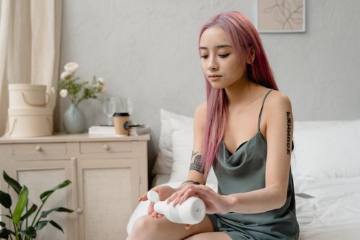 Woman with pink hair applying moisturizer in a serene bedroom setting.