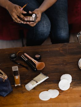 A detailed view of various makeup products on a wooden table with hands holding a palette.