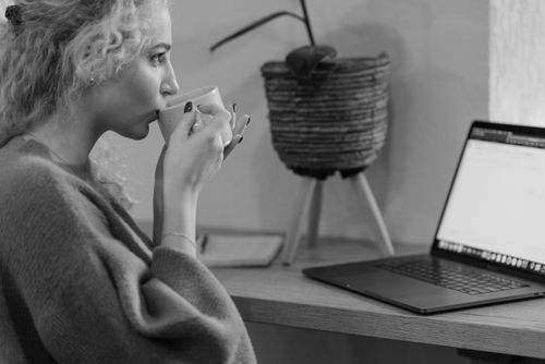 Free Black and white image of a young woman sipping a hot drink while working on a laptop indoors. Stock Photo