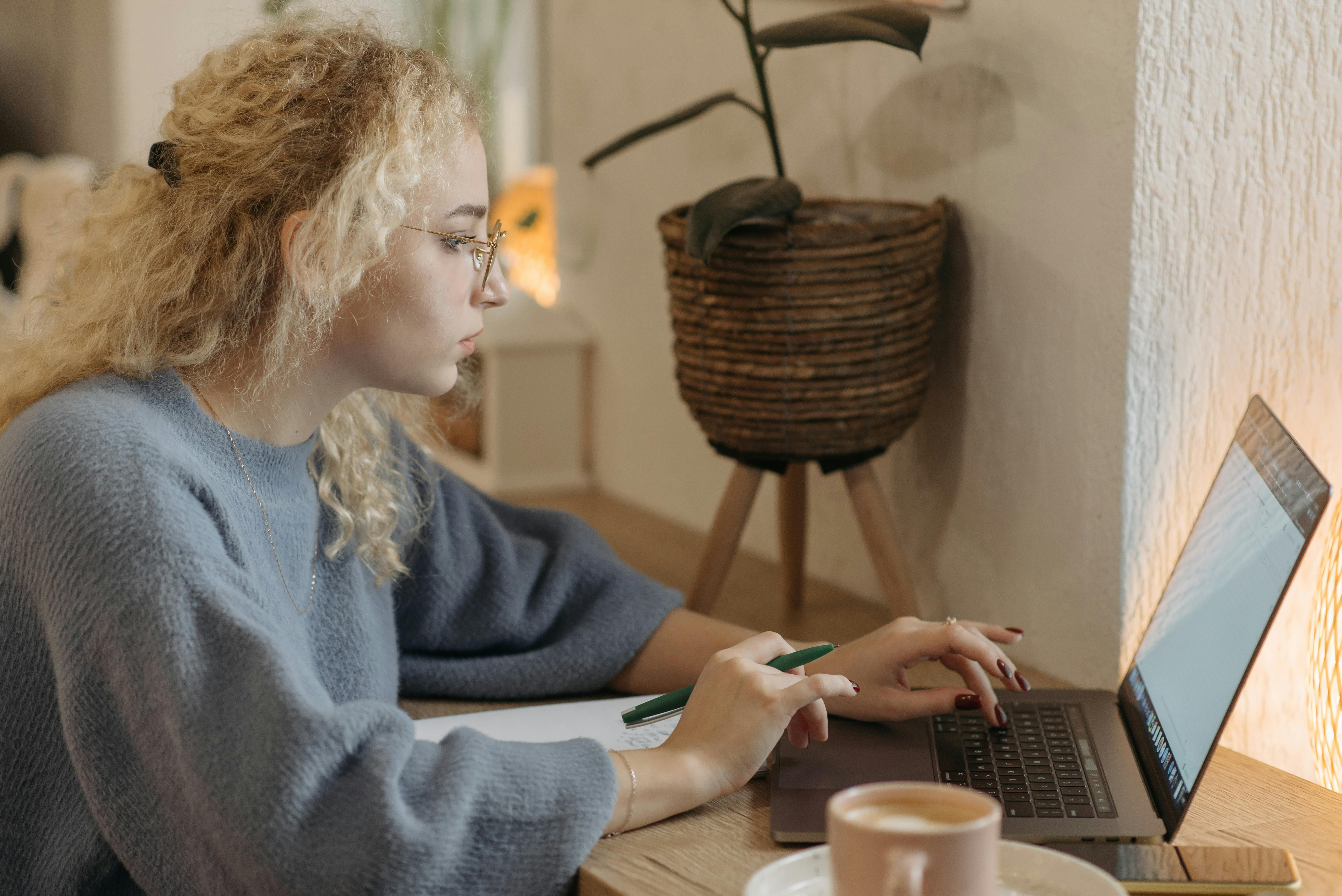 Young woman focused on remote work in a cozy indoor setting with a laptop and coffee cup.