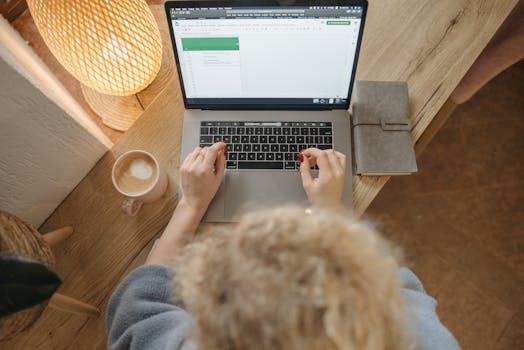Overhead view of a person working on a laptop with coffee and journal on a wooden desk.