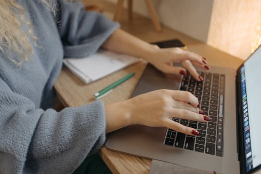 Person with red nails typing on a laptop at a wooden desk with a notebook and pencil nearby.
