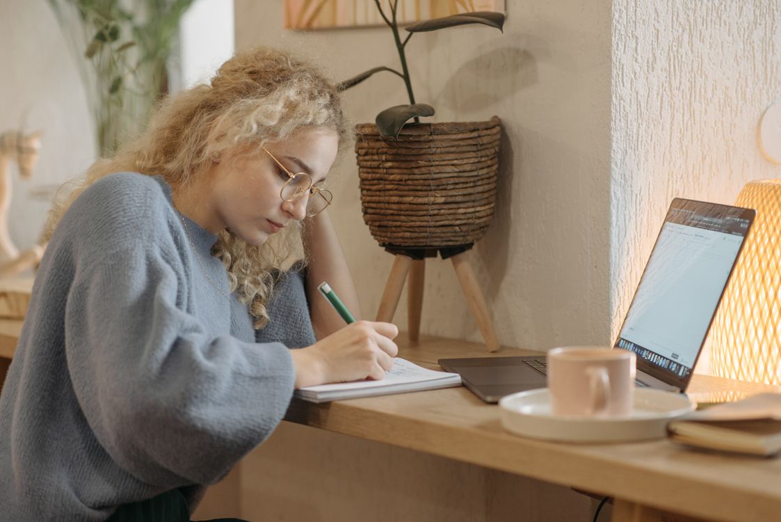 A woman writing notes at a cozy desk beside her laptop and coffee