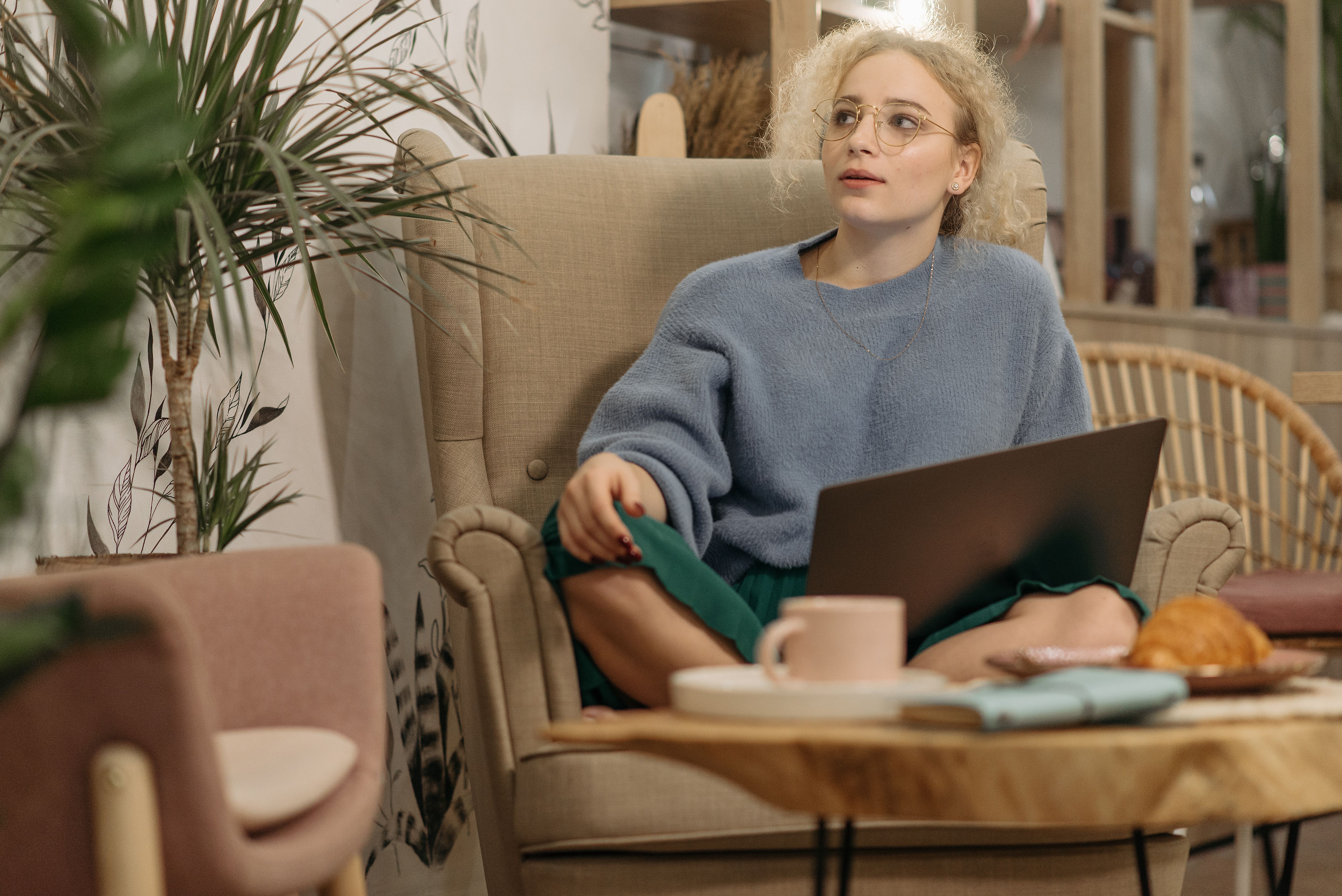 Woman in cozy setting with laptop and coffee on armchair indoors.