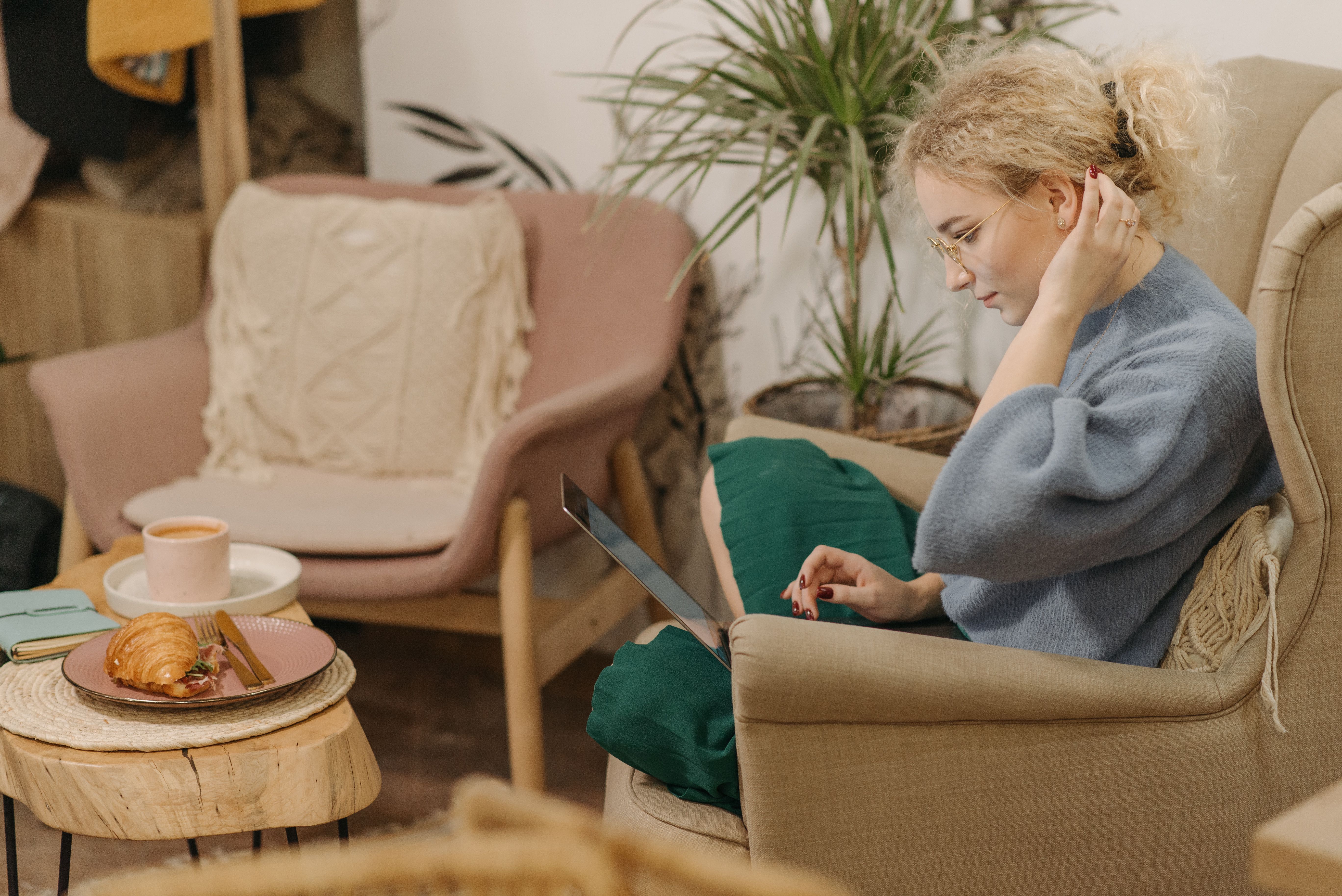 A woman sitting in a comfortable armchair working on a laptop with a coffee and croissant nearby.