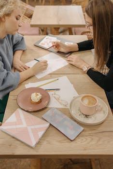 Two women collaborate on design ideas at a cozy cafe table with coffee and dessert.