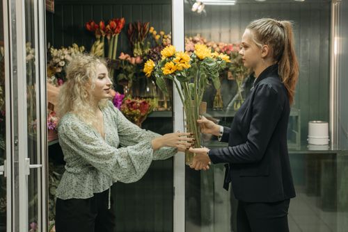 Free Two women standing, handing over sunflowers in a vibrant flower shop setting. Stock Photo