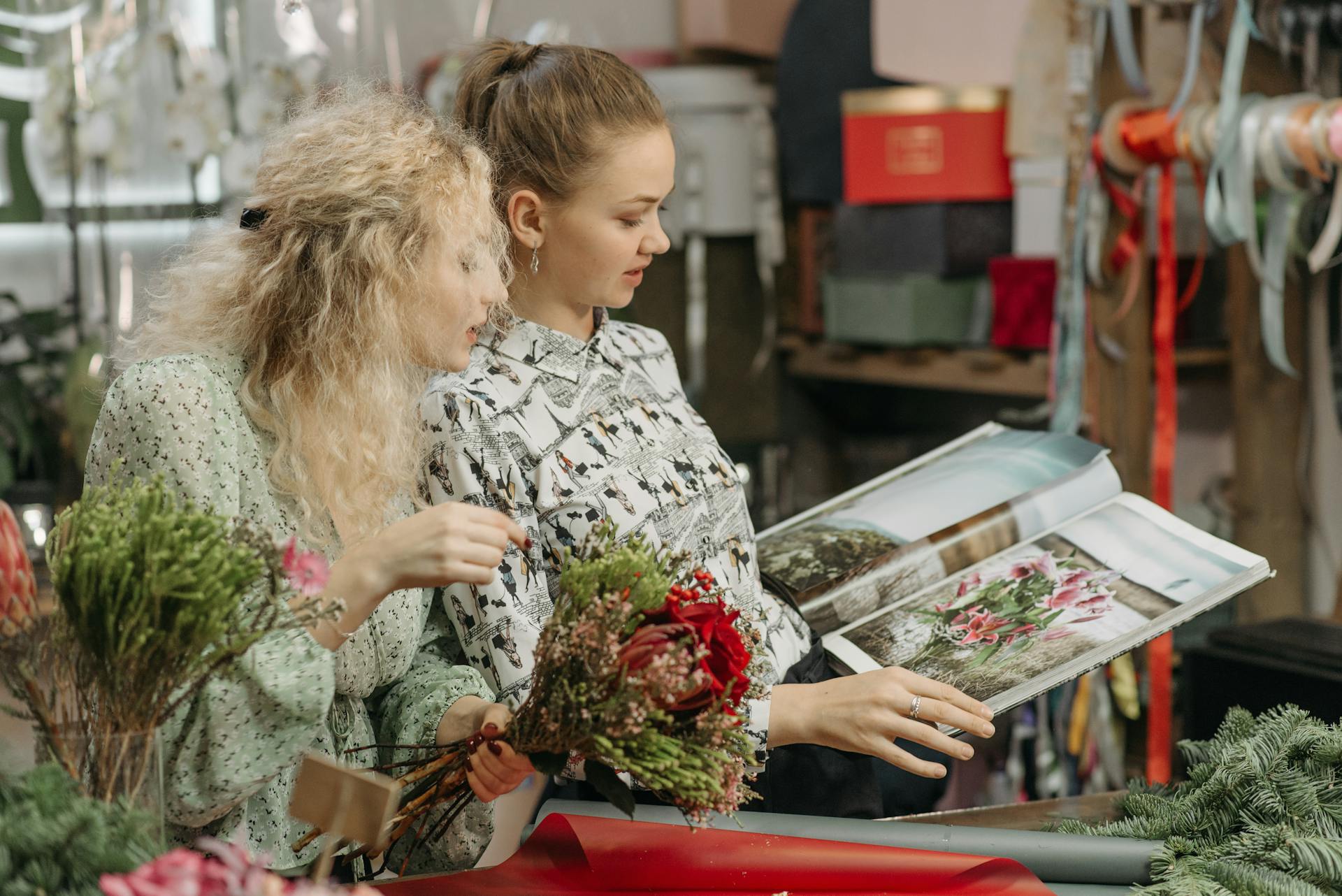 Two women florists arranging vibrant flower bouquets in a small business shop, reading a floral magazine.