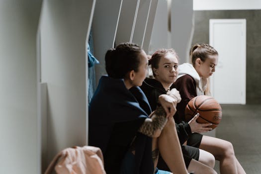 Three teenage girls sitting in a locker room, holding a basketball and chatting after practice.