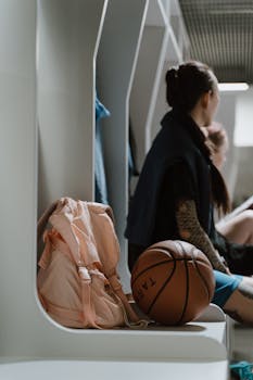 A female basketball player sitting in a locker room with a basketball and backpack.