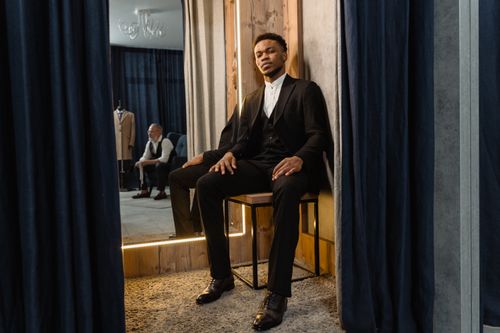 Free A stylish man in a suit sits in a tailor shop fitting room, embodying elegance and sophistication. Stock Photo