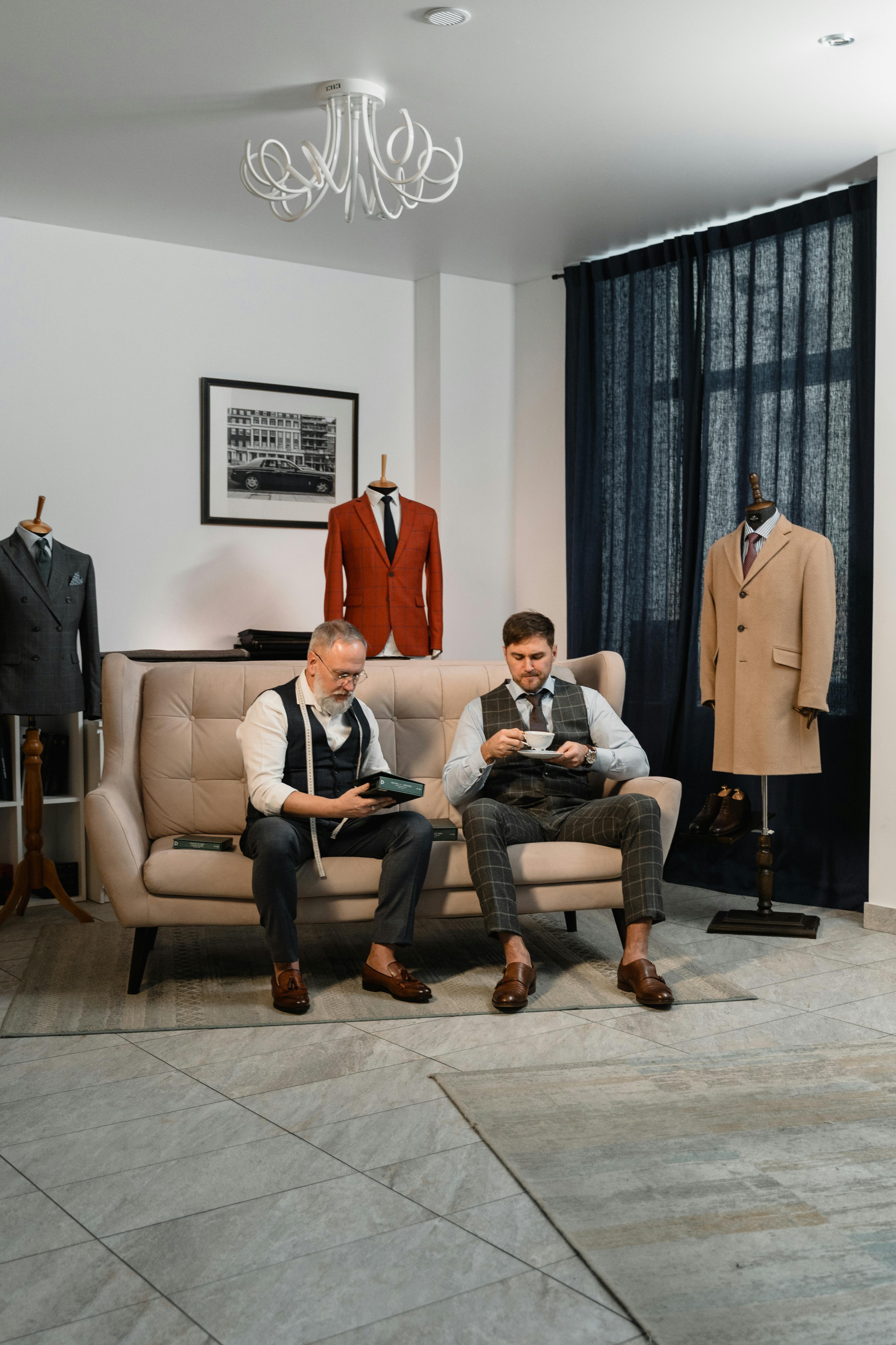 Two tailors seated in a stylish atelier chatting surrounded by elegant suits on mannequins.