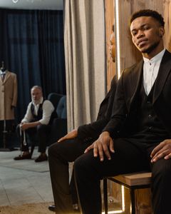 African American man in a stylish suit sitting in a modern tailor shop with a mirror reflection.