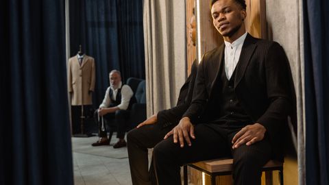 African American man in a stylish suit sitting in a modern tailor shop with a mirror reflection.