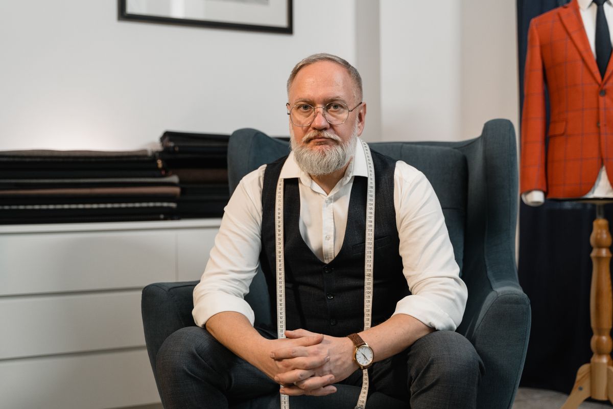 A tailor sitting in a fitting room with a tape measure around his neck