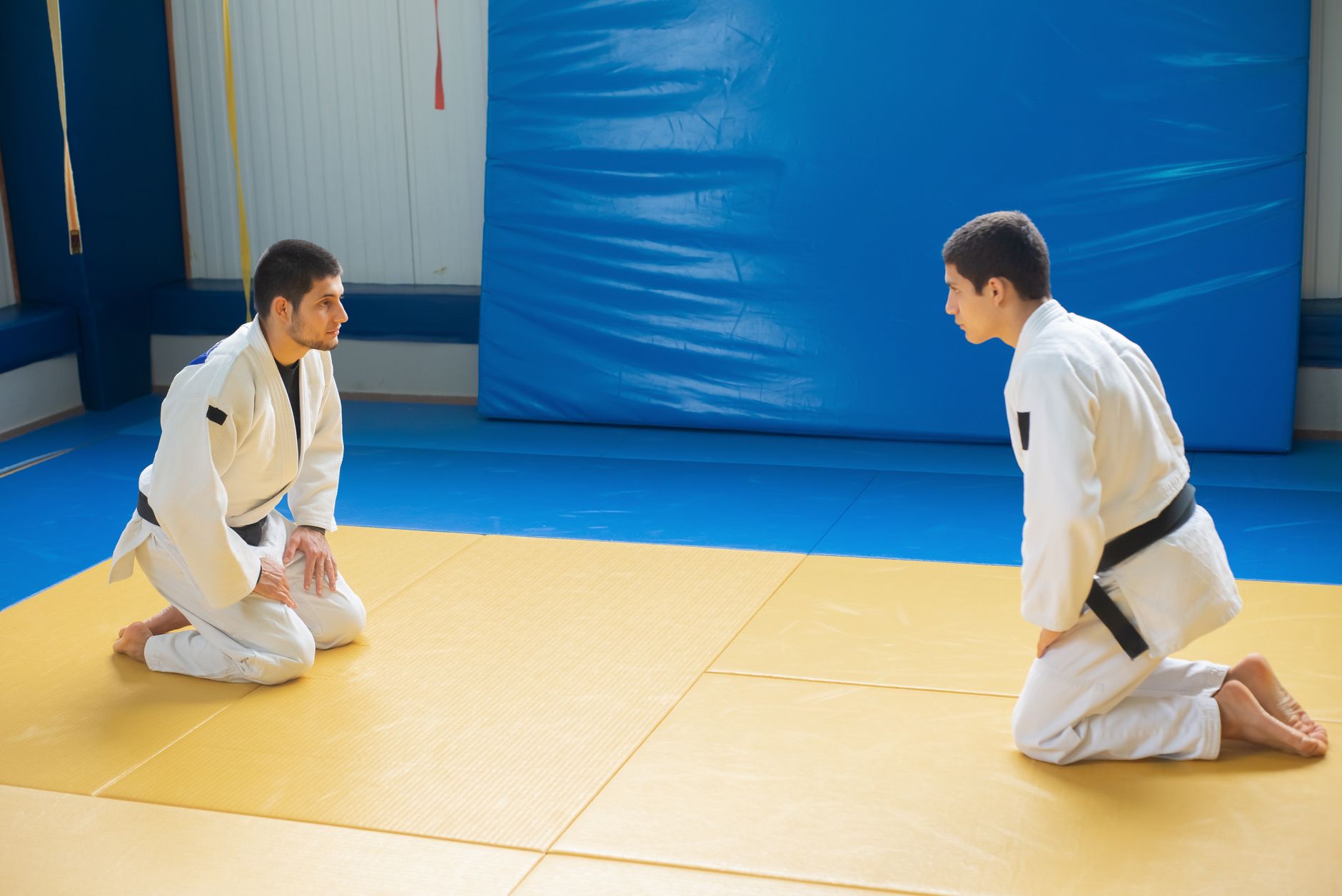Two male judoka practicing martial arts on a yellow and blue dojo mat indoors.
