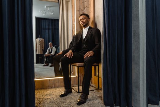 A well-dressed man in a black suit sitting confidently in a tailor shop fitting room.