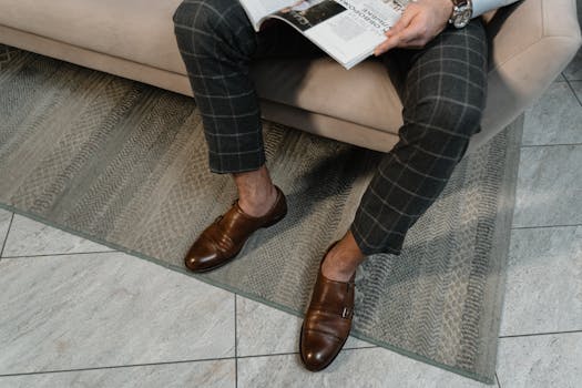 Well-dressed man in brown leather shoes and checkered pants reading a magazine indoors.
