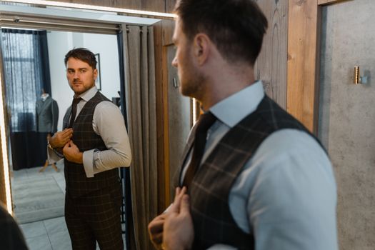 Man in a vest and shirt adjusts his suit while looking into a dressing room mirror.