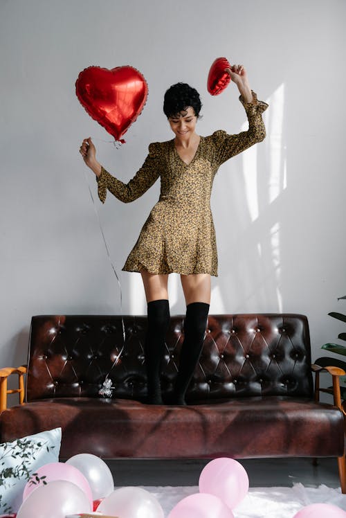 Full body of young positive female with shiny red heart shaped balloons on leather sofa