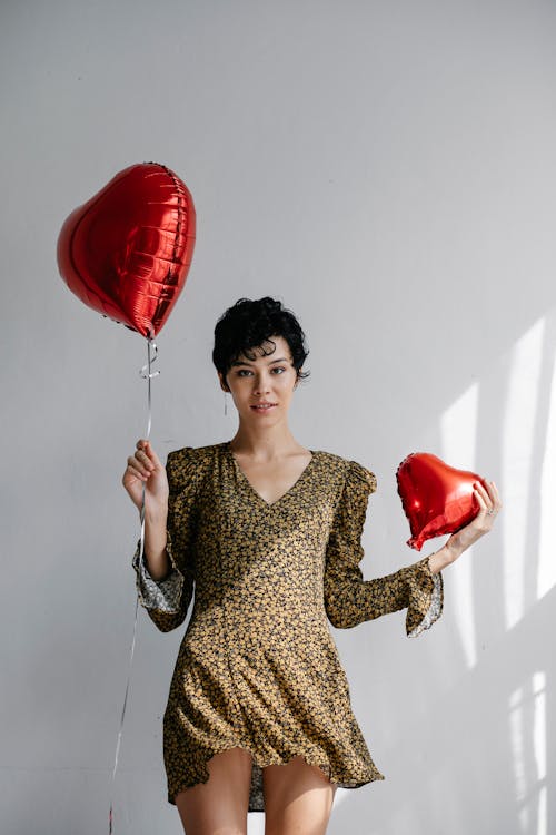 Young female with short dark hair showing bright red heart shaped balloons while celebrating Saint Valentine day