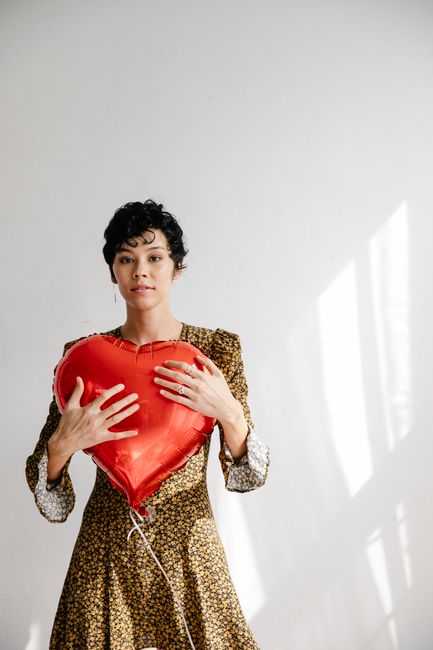 Serious female with red heart shaped balloon for Saint Valentine day standing against white wall