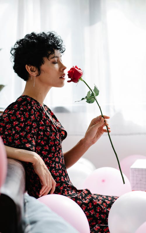 Side view of female with closed eyes smelling blooming rose while sitting on sofa with balloons for holiday celebration