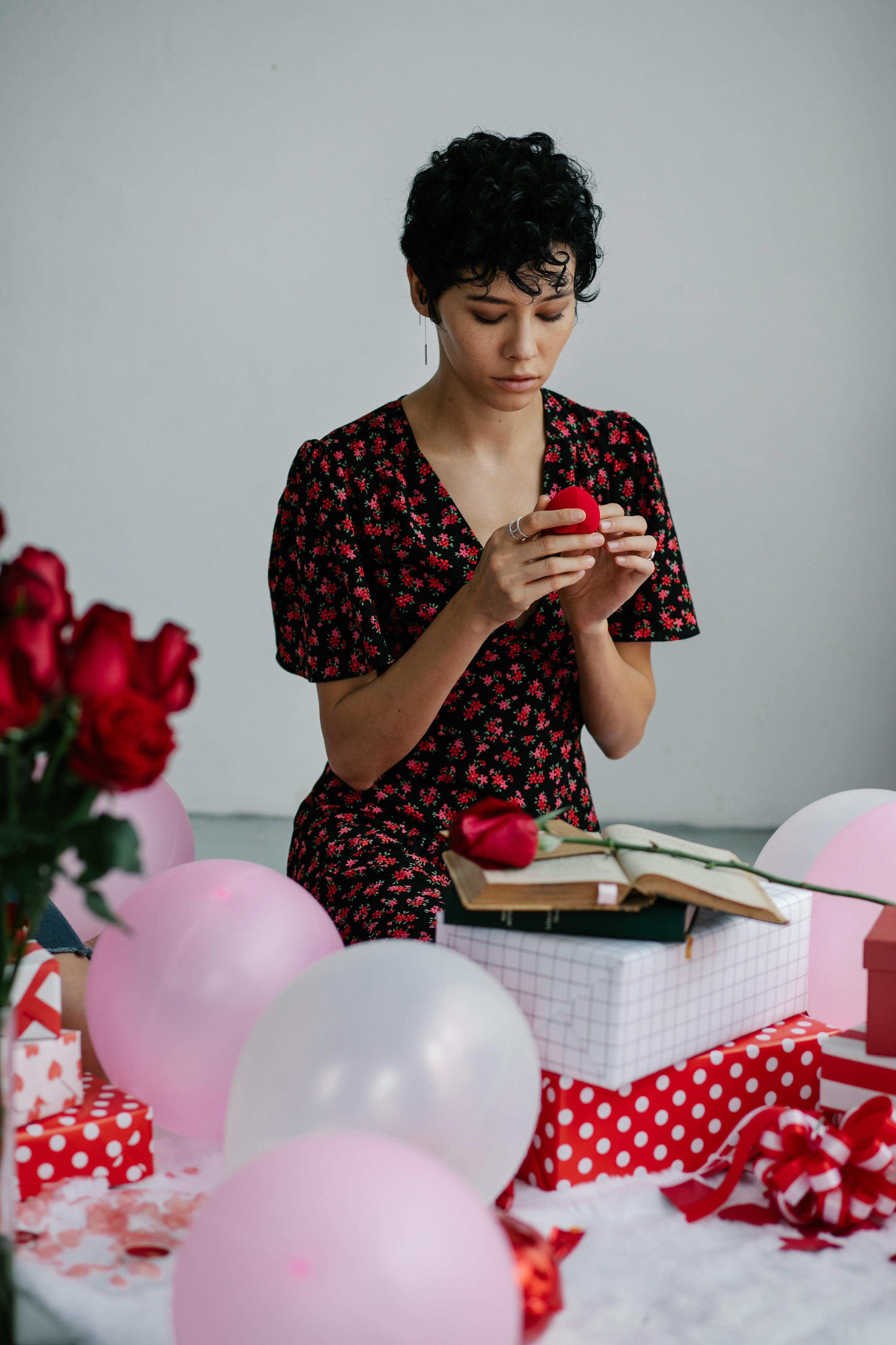 A young woman holding a gift surrounded by balloons, roses, and presents in a romantic setting.