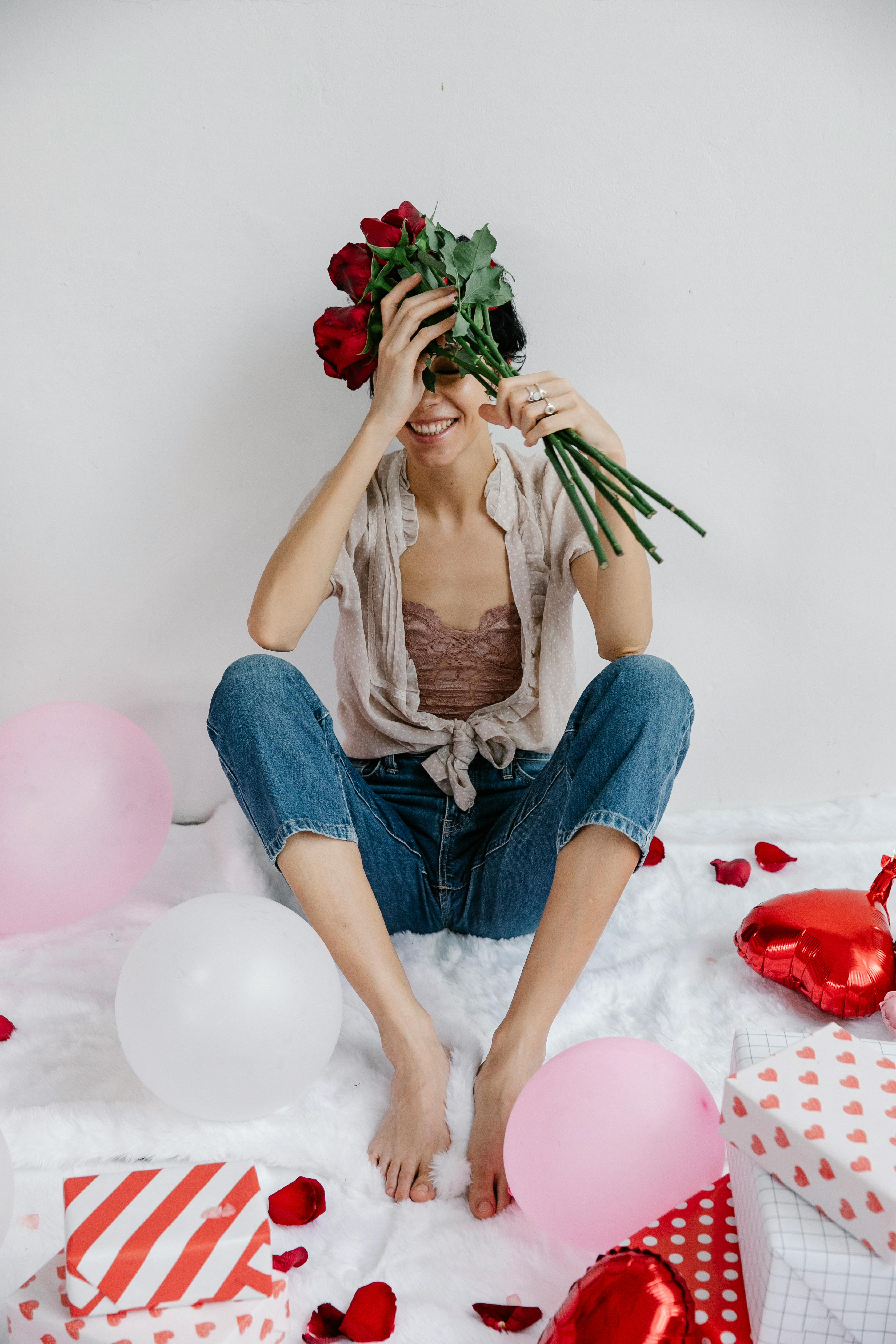 Smiling woman holding red roses surrounded by balloons and gifts, creating a festive atmosphere.