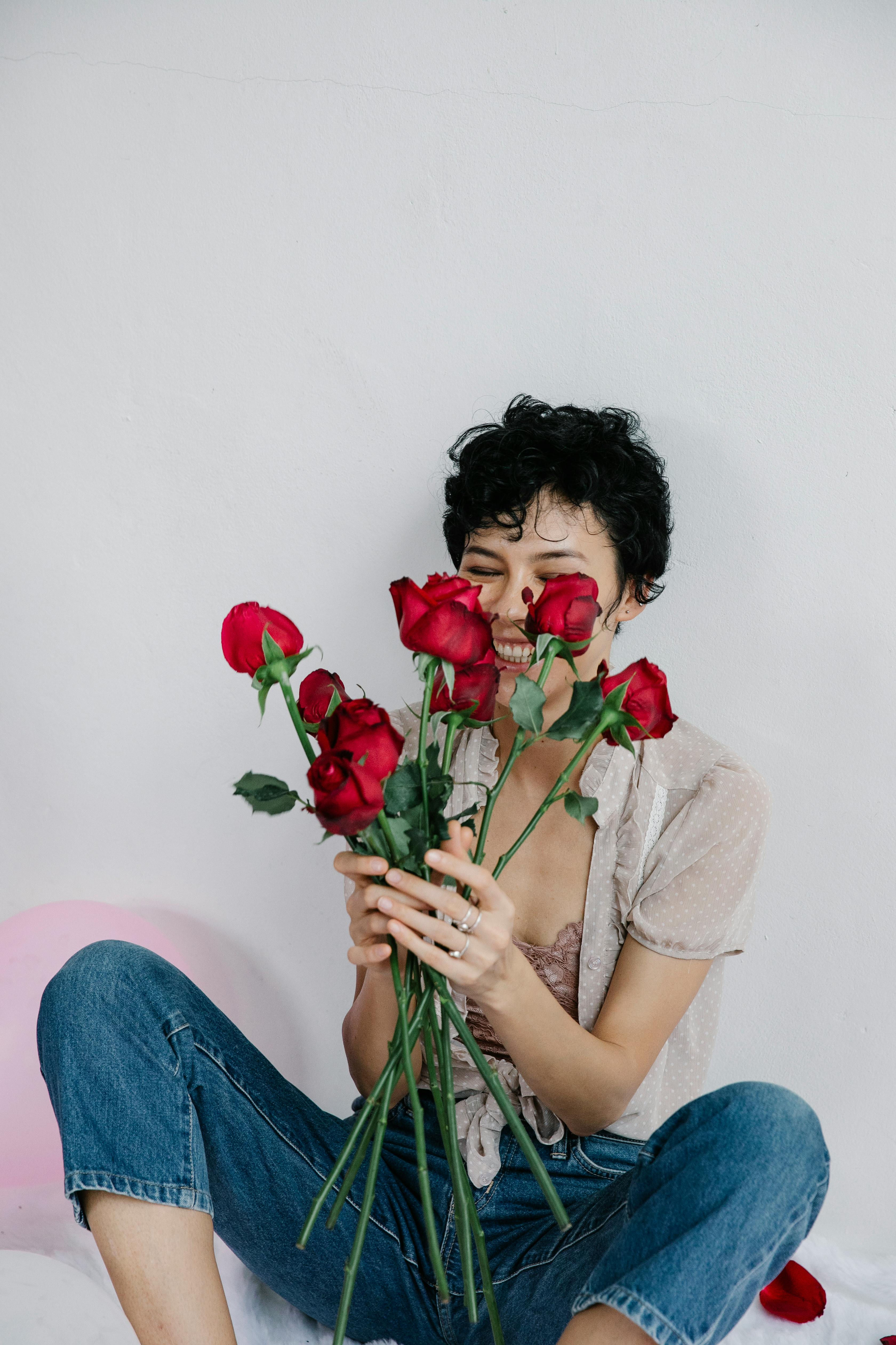 A woman with short hair smiling while holding a bouquet of vibrant red roses in a casual indoor setting.