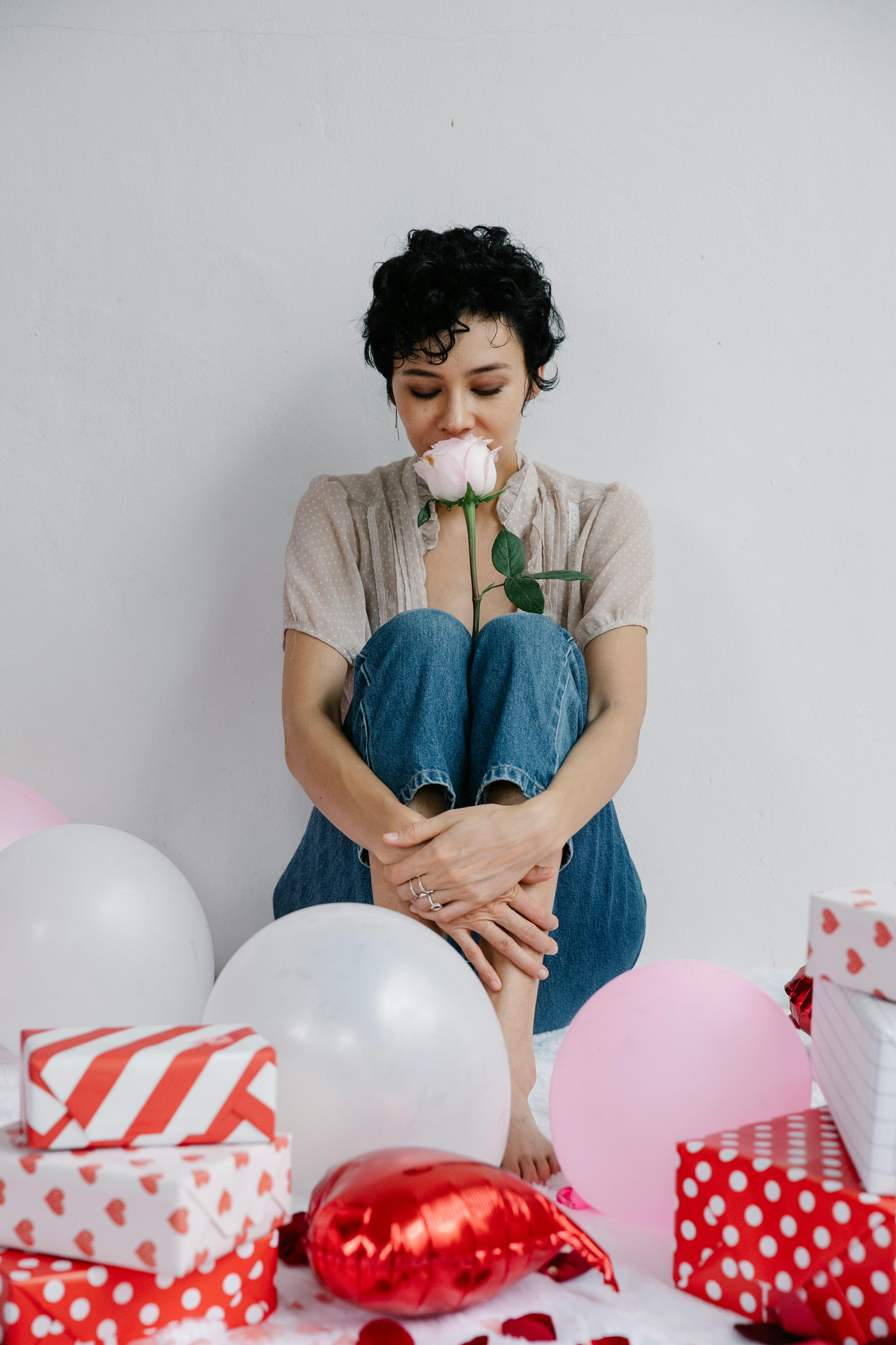 A woman sits surrounded by gifts and balloons, savoring a pink rose on Valentine's Day.