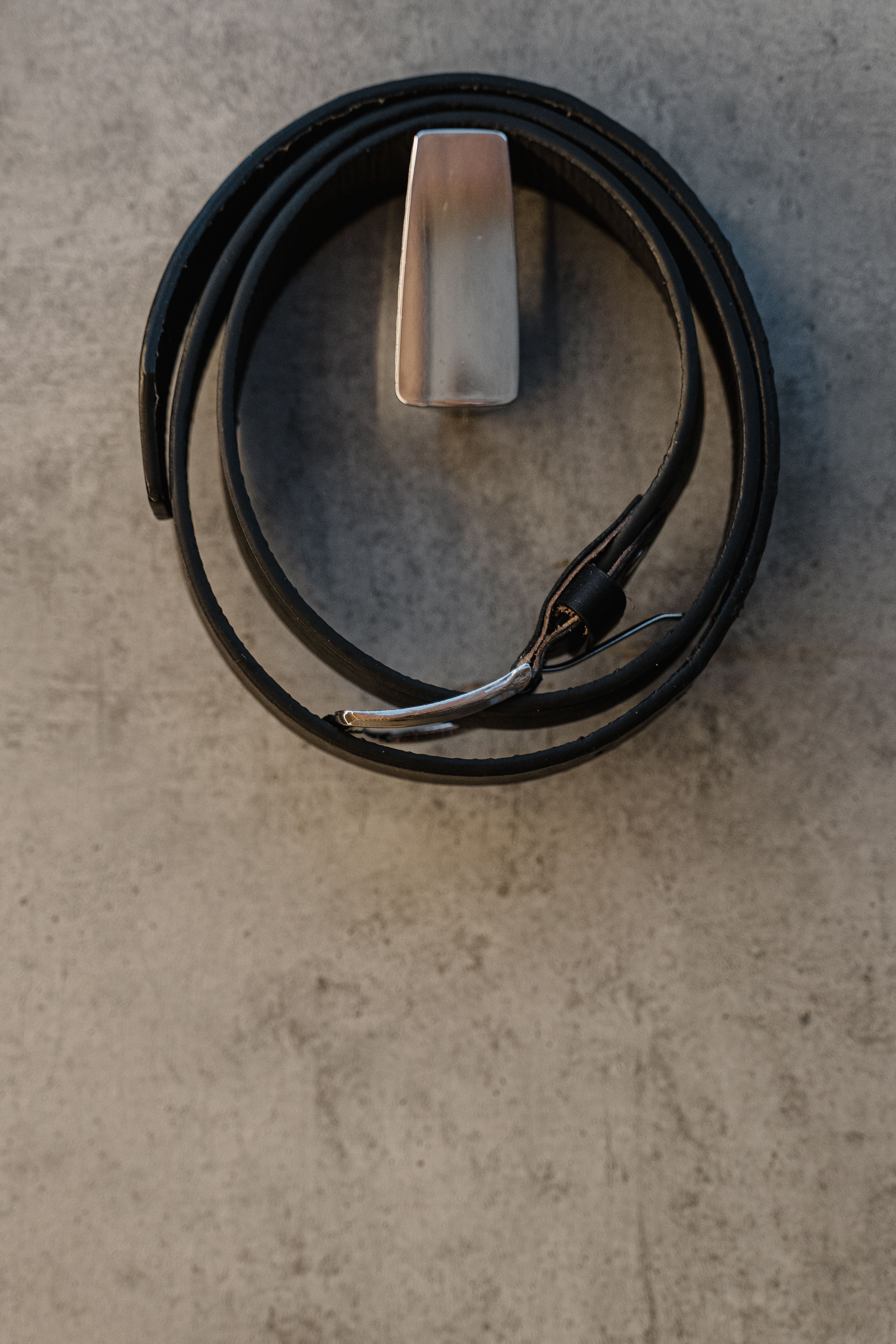 Elegante zwarte leren riem met zilveren gesp, netjes opgerold op een betonnen achtergrond voor een stijlvolle uitstraling.