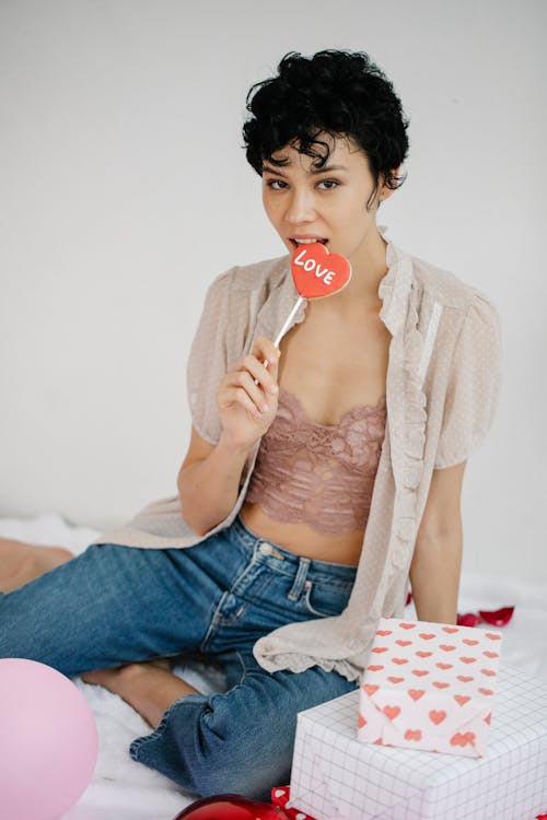 Free Attractive female eating heart shaped candy with Love inscription and looking at camera while sitting on floor with colorful balloons and gift boxes during Saint Valentines Day celebration Stock Photo