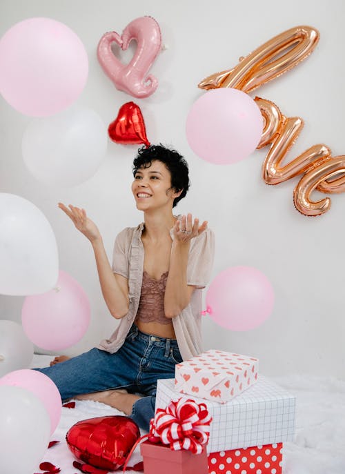 Free Cheerful female throwing up colorful balloons while sitting on floor with gift boxes near wall with Love inscription during Saint Valentines day celebration Stock Photo
