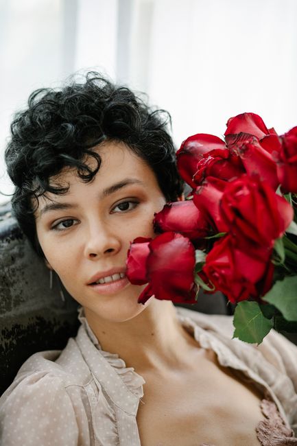 Self assured young ethnic female with dark curly hair sitting on sofa with bouquet of elegant red roses