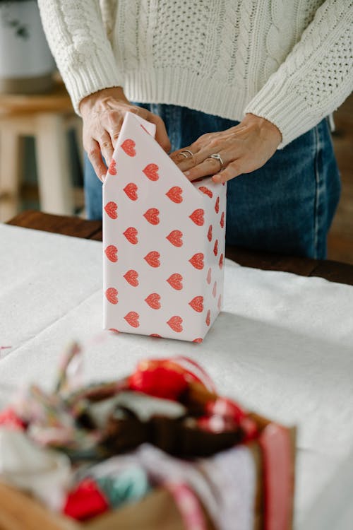 High angle of crop faceless female in knitted sweater packing gift box with wrapping paper with red hearts while standing at table at home