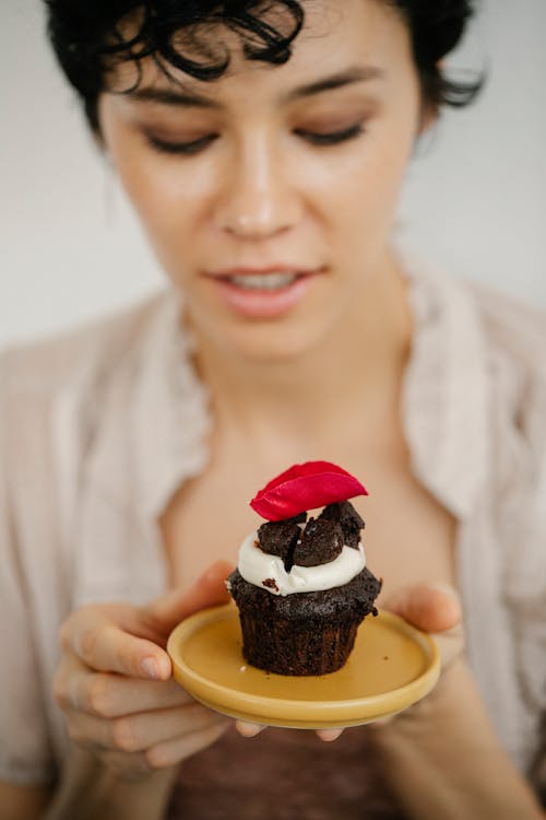 Free Delighted young ethnic lady with dark hair holding plate with appetizing chocolate muffin with whipped cream Stock Photo