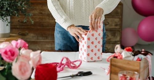 Crop anonymous lady decorating gift box with wrapping paper with red hearts while standing at table with ribbons and flowers