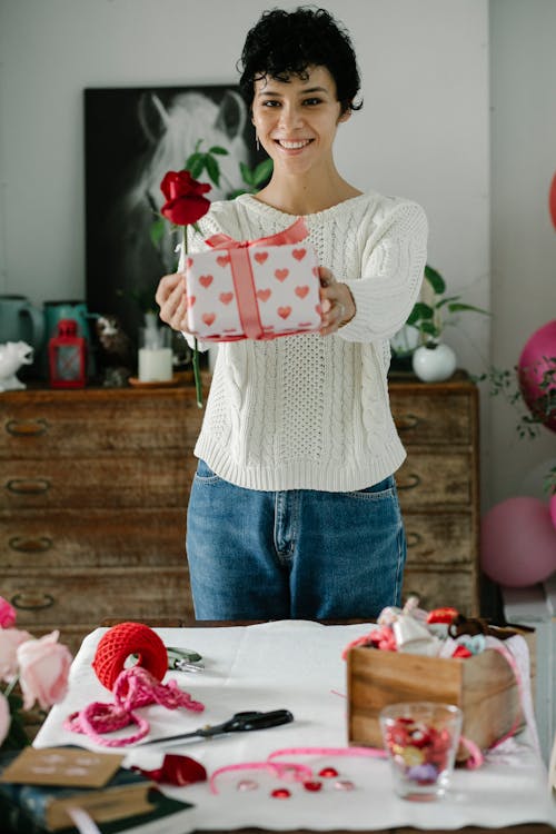 Cheerful young ethnic female with short curly hair in casual clothes smiling while demonstrating wrapped gift box and red rose and standing at table at home