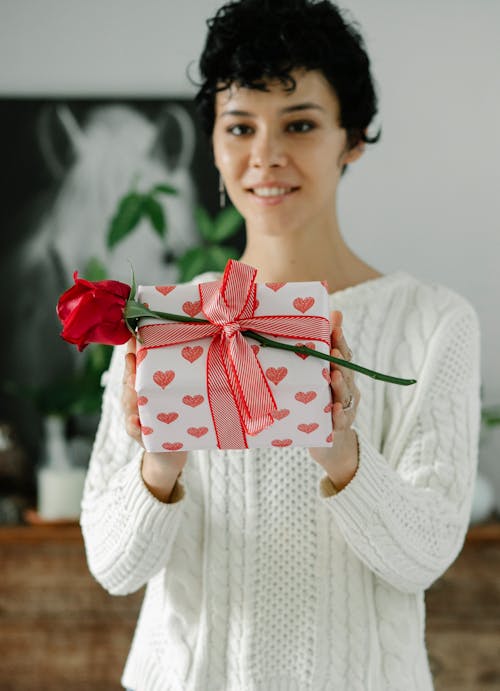 Content young ethnic female demonstrating gift box and red rose tied with ribbon