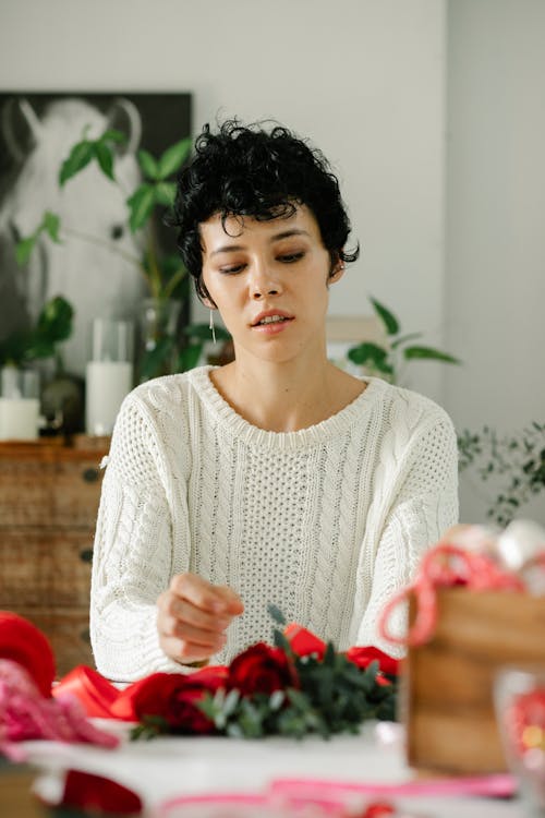 Young ethnic female florist with short curly hair in stylish clothes sitting at table and making elegant roses bouquet in light store