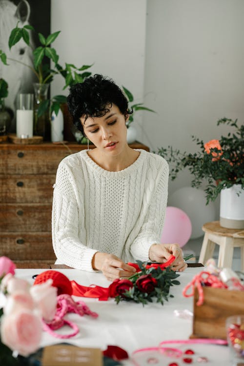 Confident young ethnic female florist with short dark hair in casual outfit tying red ribbon on bouquet of fresh roses during work in store