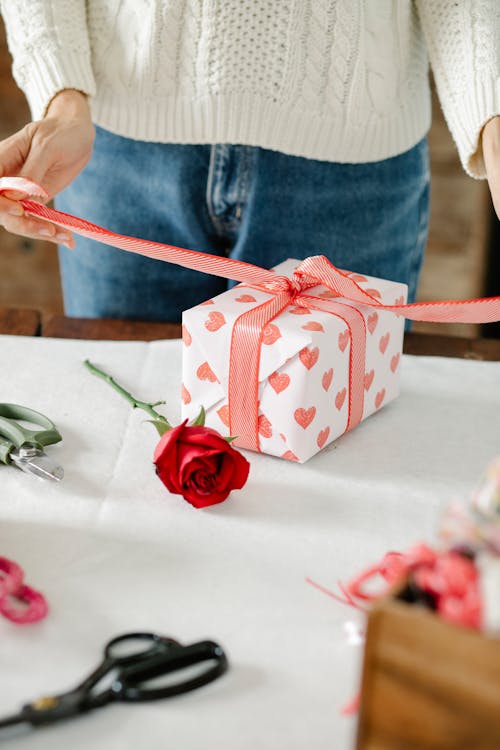 Person tying a red ribbon around a heart-patterned gift box next to a single red rose on a crafting table.