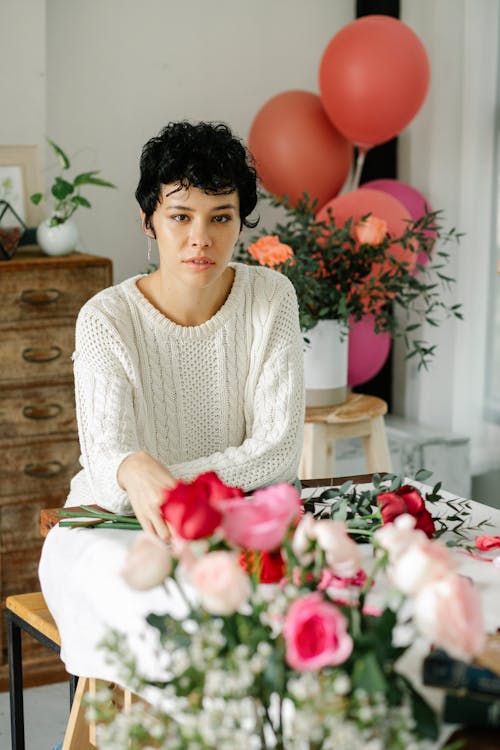 Serious young ethnic lady sitting at table with assorted flowers in decorated room