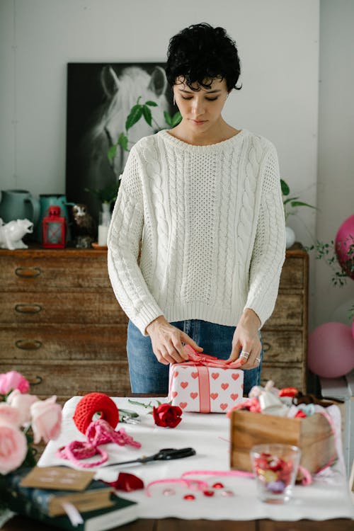 Concentrated female tying ribbon of present box while standing at table with rose and decorations supplies during festive event preparation