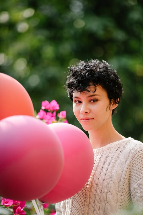 Cheerful female with short hair looking at camera while standing with bright balloons among green trees on blurred background during holiday celebration