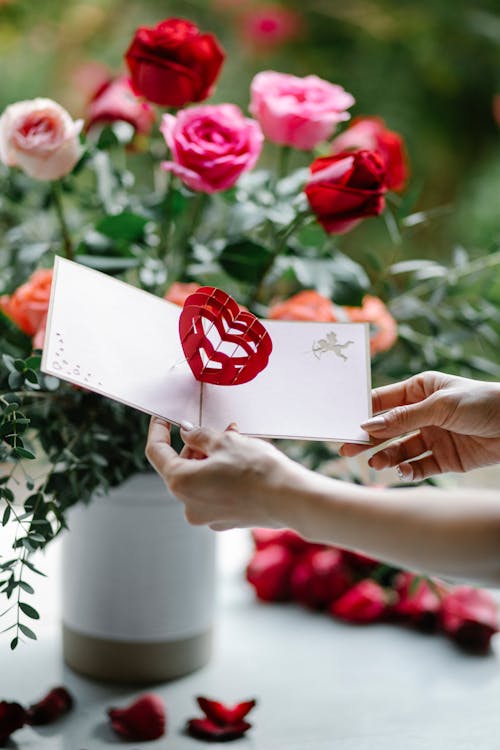 Free Unrecognizable female with postcard with red 3d heart standing near table with bouquet of roses in vase on blurred background Stock Photo