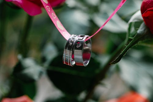 Closeup of rings with engraved Love inscription hanging on pink ribbon near colorful flowers with green stems on blurred background during romantic holiday