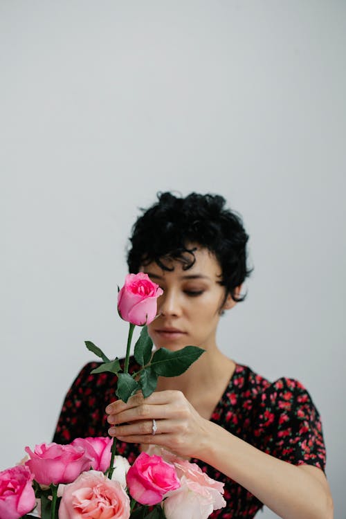 Serious female with short hair putting pink rose in bunch of fresh flowers while standing near white background during holiday